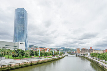 Obraz premium A view of the embankment in the city of Bilbao, Spain, from the Nervion River A large, tall, blue skyscraper is located on the embankment. The weather is cloudy, with an overcast sky