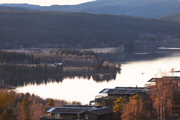 Evening light reflects over calm Norwegian lake near Oslo, mirroring dark pine forests and distant houses in tranquil Nordic autumn atmosphere
