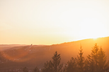 Holmenkollen ski jump rises through golden haze above Oslo pine-covered hills during sunset, glowing in warm Scandinavian spring atmosphere