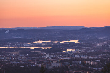 Golden rays touch treetops above Oslo as sun sets over hills and fjord, creating a warm balance between Nordic wilderness and urban horizon. Grefsenkollen