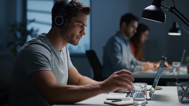 Focused male programmer with headphones working late on a laptop in a modern office, deeply concentrated and typing code while staying hydrated during a late night freelance session