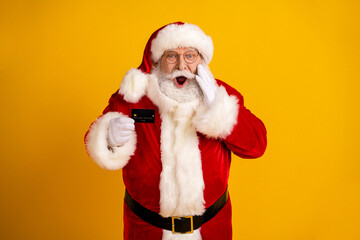 Surprised Santa holds card in red suit with white fur against bright yellow background for Christmas themed stock photo