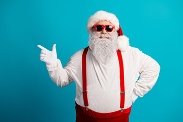 Santa in red suspenders and hat with white beard and sunglasses points against blue background in a...