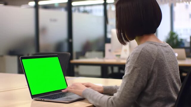 A focused young woman with short dark hair works diligently on a laptop with a green screen in a modern openplan office environment during the day showcasing remote work and digital tasks.