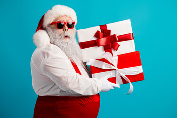 Santa Claus in red suit with white gloves carrying stacked gift boxes against a blue background for christmas shopping and holiday season stock photo