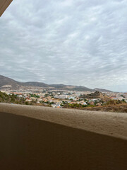 Panoramic view of a mountain landscape and a town in a valley, seen from behind a balcony railing. Spain, 2022.