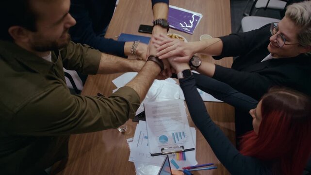 Office workers unite by putting their hands together in a gesture of teamwork and collaboration. The top view shows a wooden table with financial documents and some snacks beneath