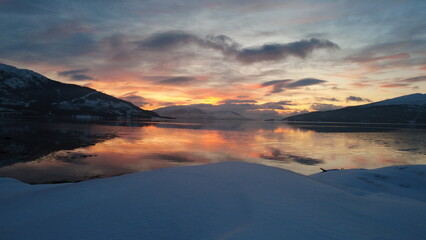 A breathtaking drone shot of a sunset illuminating a snowy fjord, surrounded by majestic snow-covered mountains. Vibrant orange hues dance across the sky behind the fjord.