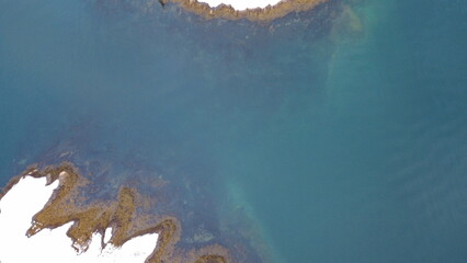 Drone photo of a rocky shoreline covered with algae, gently sloping into clear turquoise-green water, showing underwater textures and natural beauty.