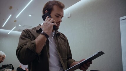 Confident businessman multitasking by holding business papers and talking on his smartphone during a workday meeting. The scene captures teamwork and concentration inside a modern corporate office - Powered by Adobe