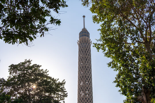 View of the Cairo Tower pierces the sky, framed by the lush green foliage of surrounding trees under a bright sun, Zamalek, Cairo Governorate, Egypt.