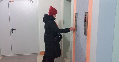 A woman presses an elevator button while waiting in the hallway in winter clothes.