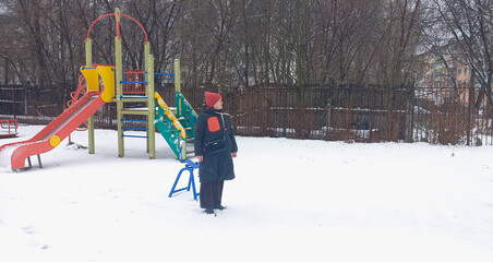 A woman in a black down jacket stands alone on a snowy playground with a colorful slide.