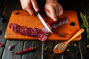 A person slices a sausage on a wooden board, surrounded by various herbs and spices. The setting is a cozy kitchen with dark wood elements, capturing the essence of cooking preparation