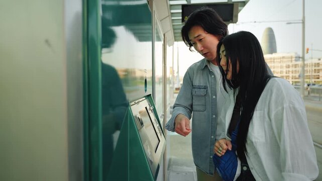 Two young Asian friends using ticket machine at tram station to buy public transport commuter travel card