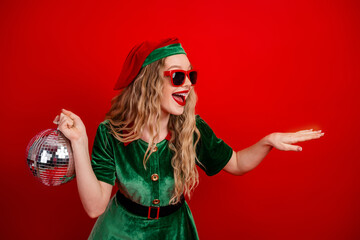 Festive young woman dressed as an elf holding a shiny disco ball and striking a playful pose with...