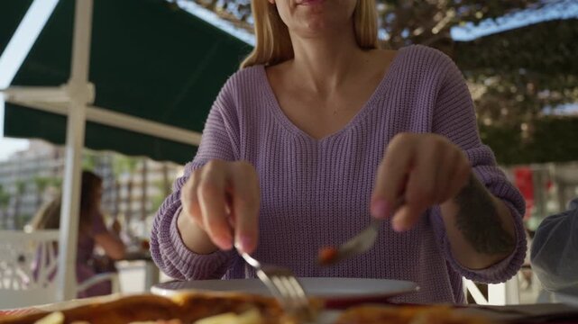 Blond woman with glasses  cut slice of pizza with knife and fork at an outdoor cafe table, enjoy sunny afternoon break from day