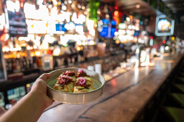A hand holds a plate of appetizers topped with pulled pork beef and pickled red onions in a lively, blurred bar or restaurant setting.
