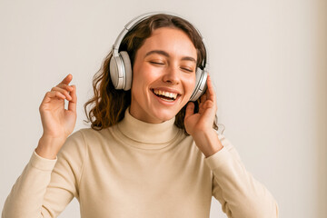 Horizontal format portrait of a cheerful young woman in a beige turtleneck dancing joyfully while listening to music with silver wireless headphones against a bright neutral background