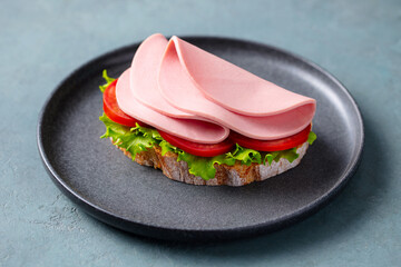 Boiled sausages sandwich with tomatoes, green salad on a black plate. Grey background. Close up.