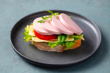 Open sandwich with boiled sausage, tomato, cheese and lettuce on rustic bread. Grey background. Close up.