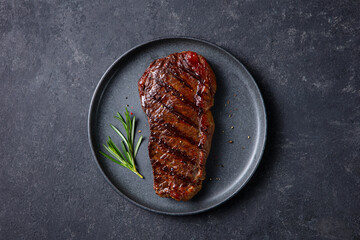 Grilled steak served on dark plate, enhanced with rosemary touch. Dark grey background. Close up. Top view.