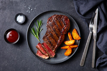 Beef Steak medium rare with roasted potato wedges on a black plate. Grey background. Close up. Top view.