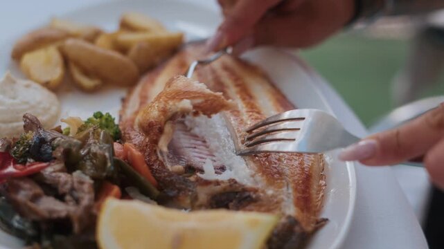 Close-up of hands cutting into baked hake fish with fork and knife, roasted potatoes, and vegetables