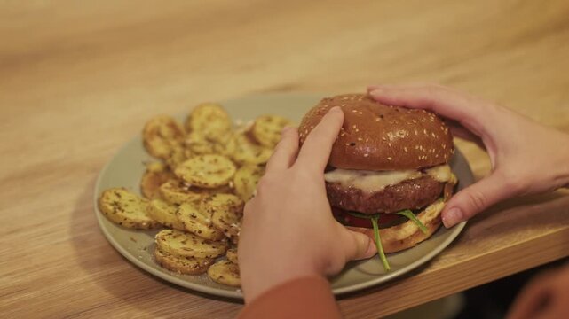Woman&rsquo;s hands grabbing cheeseburger with French fries on plate, close-up
