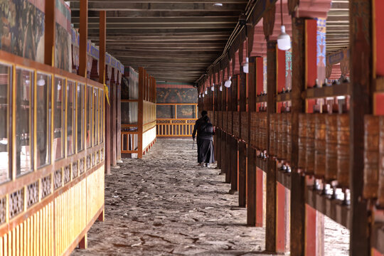 View of a lone figure walking through a long corridor with wooden pillars and stone floors, ancient murals adorning the walls, Shannan, China.