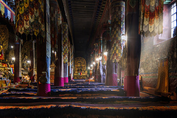View of a dimly lit temple hall with ornate columns wrapped in magenta fabric, leading to a golden Buddha statue, Shigatse, Tibet, China.