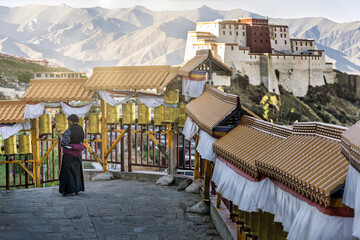 View of a woman in traditional dress, standing near prayer wheels and overlooking the imposing fortress against a mountainous backdrop, Shigatse, Tibet, China.
