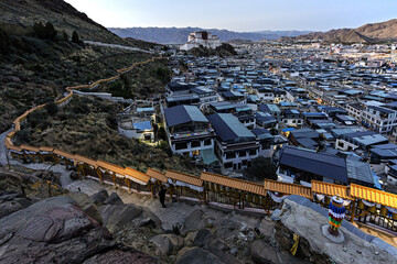 View of a winding stone path lined with prayer wheels descending a rugged hillside towards a dense cityscape under a vast sky, Shigatse, Tibet, China.