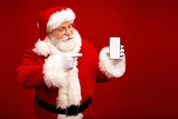 Santa holds a smartphone and points at it against a rich red background in a festive portrait of holiday cheer and modern tech