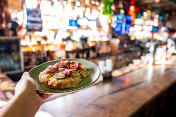 A hand holds a small green plate featuring a personal-sized flatbread pizza topped with cheese, bacon, and pickled red onions, set against a blurry, warm-toned bar background