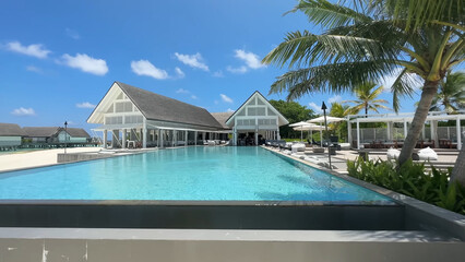 Luxury resort photo of Maldives tropical paradise island in Indian Ocean with swimming pool, palm trees, beach umbrella, chair. Beautiful photography, wooden house, sky with clouds.