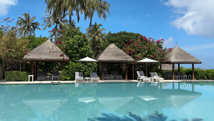 Luxury resort photo of Maldives tropical paradise island in Indian Ocean with swimming pool, palm trees, beach umbrella, chair. Beautiful photography, wooden house, sky with clouds.