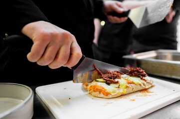 Close-up of a chef in black uniform slicing a melted, loaded, open-face sandwich on a white cutting board.