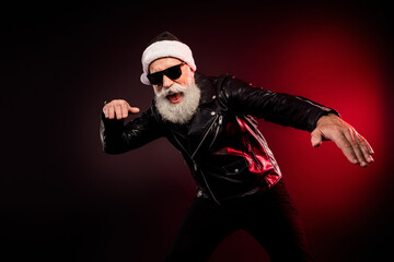 Festive rocker santa in a leather jacket dancing with a white beard and sunglass under red light for christmas promotion and holiday party vibes