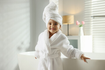 Cute girl in bathrobe with wrapped hair near bathtub indoors