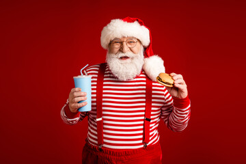 Santa in striped shirt with red suspenders and hat holds a burger and a blue cup against a solid...