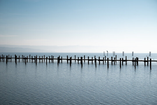 View of weathered wooden posts standing starkly against the tranquil water, reaching towards the horizon under a serene sky, Schondorf am Ammersee, Bavaria, Germany.