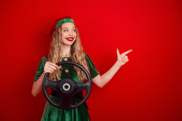 Festive elf in green holiday attire holding a steering wheel against a red background cheerfully...
