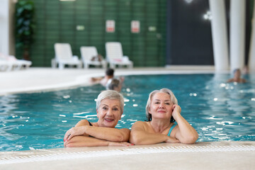 Two smiling senior women enjoying leisure time in an indoor swimming pool, leaning on the poolside. Concept of friendship, wellness, active lifestyle, relaxation, and healthy aging.