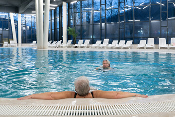Two senior women enjoying relaxation while leaning on the edge of a modern indoor swimming pool in the evening. Concept of wellness, healthy lifestyle, spa, leisure, and friendship for older adults.