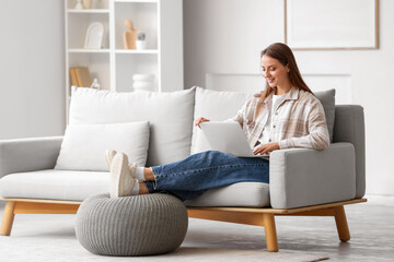 Obraz premium Young woman using laptop with pouf on sofa in living room