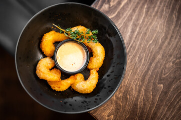 breaded shrimp arranged in a circle around a small bowl of creamy dipping sauce, garnished with thyme, served in a black bowl on a dark wooden table.
