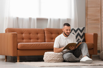 Obraz premium Young man with pouf reading magazine on carpet in living room