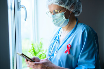 A weary female doctor with red awareness ribbon on uniform checks her phone, maintaining connection with HIV AIDS patients during work.