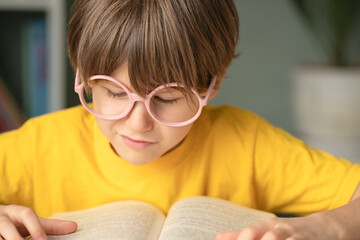 Smiling pupil in glasses rests hand while reading open book at school desk. Childhood learning, clever mind, study concept, early development and preparation for education and future growth.